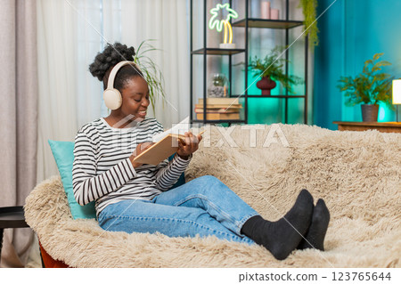 African American woman young adult reading novel, deeply focused, sitting on sofa, relaxing at home 123765644
