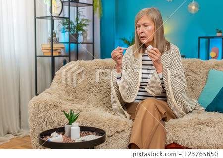 Mature woman covered with blanket on sofa checking temperature with thermometer wipes snot at home Mature woman covered with blanket on sofa checking temperature with thermometer wipes snot at home 123765650