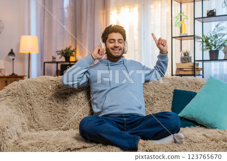Indian young man sitting on sofa listening to music with headphones dancing to rhythm at home Indian young man sitting on sofa listening to music with headphones dancing to rhythm at home 123765670