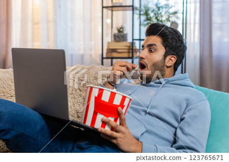 Indian young man eating popcorn and watching movie on laptop while sitting on sofa in living room Indian young man eating popcorn and watching movie on laptop while sitting on sofa in living room 123765671