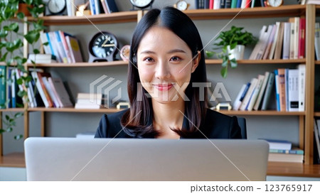 Asian woman working in financial office, Asian woman reviewing documents, laptop open. Financial office featuring bookshelves, clocks, reflecting business organization, financial expertise concept. 123765917