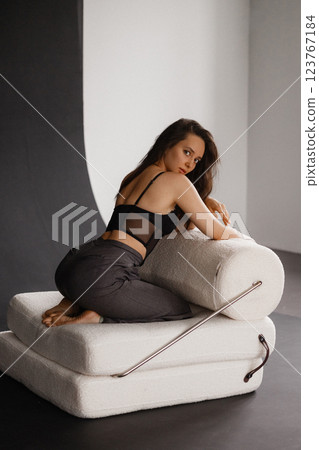Stylish woman in black corset and grey trousers on modern white sofa. Back view. 123767184