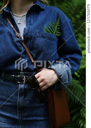 Close-up of a stylish woman wearing a denim outfit with a casual and natural aesthetic. A green fern leaf is tucked into the chest pocket, adding a fresh and eco-friendly touch 123768079