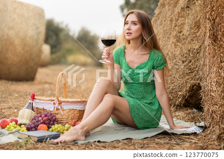 Young beautiful woman in summer green dress sitting and drinking on soft mat near stack of straw with glass of wine, romantic picnic in field Young beautiful woman in summer green dress sitting and drinking on soft mat near stack of straw with glass of wine, romantic picnic in field 123770075