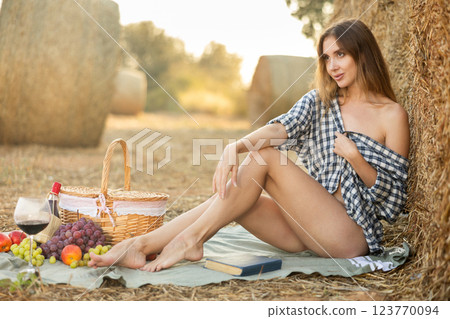 Beautiful cowboy woman in checked shirt sitting and posing near haystacks with basket of fruit and wine, fashion concept Beautiful cowboy woman in checked shirt sitting and posing near haystacks with basket of fruit and wine, fashion concept 123770094