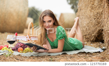 Young smiling sexy woman wearing in summer green dress lying down and reading book on soft mat near with stack of straw and enjoying romantic picnic 123770169