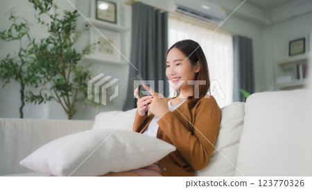 Happy young asian woman relax on comfortable couch at home texting messaging on smartphone Happy young asian woman relax on comfortable couch at home texting messaging on smartphone 123770326
