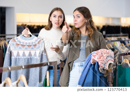 Amazed female holding warm clothes on hangers and showing her friend a discount during shopping in a store Amazed female holding warm clothes on hangers and showing her friend a discount during shopping in a store 123770413