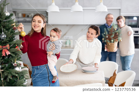Young woman with a baby in her arms decorates a Christmas tree with toys while the rest of the family prepares festive dinner Young woman with a baby in her arms decorates a Christmas tree with toys while the rest of the family prepares festive dinner 123770536