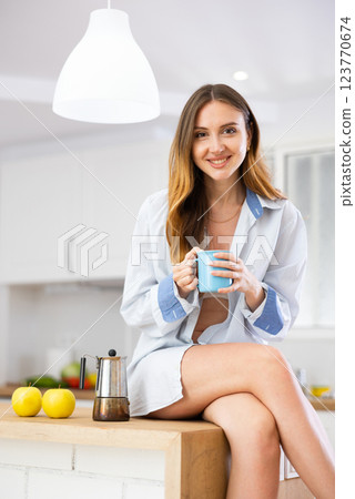 Portrait of happy young woman in shirt drinking morning coffee in modern kitchen Portrait of happy young woman in shirt drinking morning coffee in modern kitchen 123770674