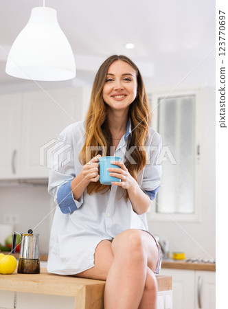 Portrait of happy young woman in shirt drinking morning coffee in modern kitchen Portrait of happy young woman in shirt drinking morning coffee in modern kitchen 123770697