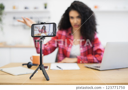 African woman sitting at desk in casual clothes recording video using smartphone tripod. 123771750