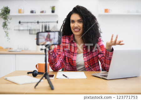 Young African woman with curly hair recording video blog in modern kitchen setup. Person wears red plaid shirt, communicates energetically on camera, and utilizes laptop for content creation. Young African woman with curly hair recording video blog in modern kitchen setup. Person wears red plaid shirt, communicates energetically on camera, and utilizes laptop for content creation. 123771753
