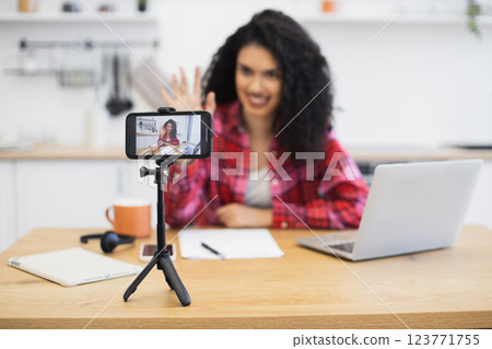 Young African woman filming content with smartphone, laptop, and stationery in kitchen setting. Creates social media content using technology at home. Young African woman filming content with smartphone, laptop, and stationery in kitchen setting. Creates social media content using technology at home. 123771755