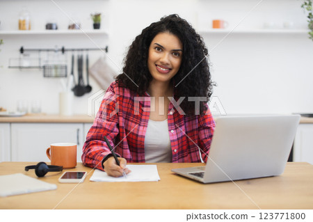 African American woman in casual attire working on computer at kitchen desk writing notes African American woman in casual attire working on computer at kitchen desk writing notes 123771800