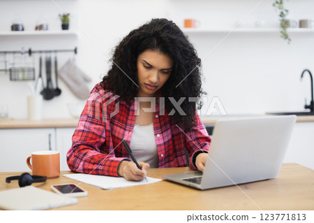 Young African American woman writing on paper while working from home using a laptop. Casual lifestyle setting with a coffee cup nearby. Creative, focused, and productive atmosphere indoor. Young African American woman writing on paper while working from home using a laptop. Casual lifestyle setting with a coffee cup nearby. Creative, focused, and productive atmosphere indoor. 123771813