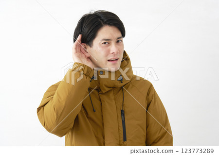 一位身穿羽絨外套的男子用手摀住耳朵聆聽，白色背景 123773289