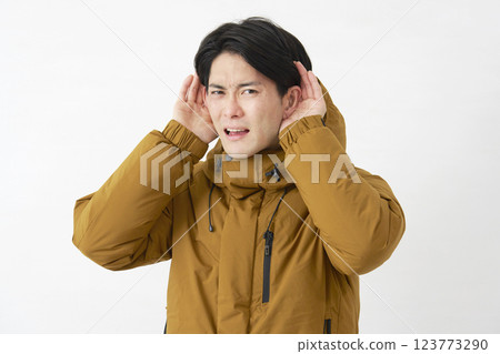 一位身穿羽絨外套的男子用手摀住耳朵聆聽，白色背景 123773290