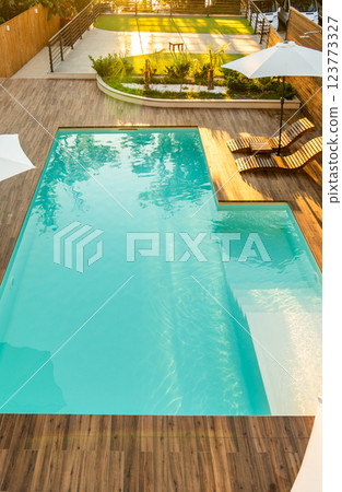 Luxurious turquoise swimming pool with wooden deck, umbrellas, and lounge chairs in a serene backyard setting 123773327