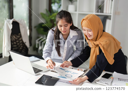 Diverse businesswomen collaborating over financial reports and laptop in a modern office, discussing strategy. 123774570