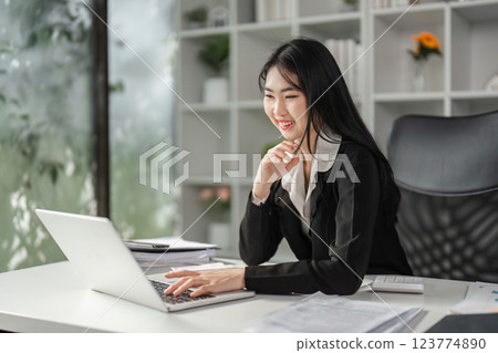 Businesswoman smiling while working on her laptop, showcasing a positive and productive work environment. Businesswoman smiling while working on her laptop, showcasing a positive and productive work environment. 123774890
