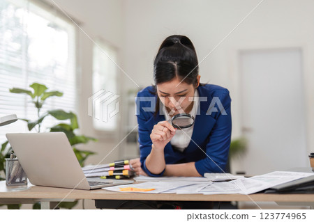 Accountant scrutinizing financial papers with a magnifying glass in a contemporary workspace. Accountant scrutinizing financial papers with a magnifying glass in a contemporary workspace. 123774965