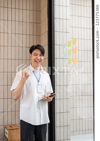Young professional celebrating success while holding tablet, standing next to organized sticky notes on glass wall in modern office. 123775366
