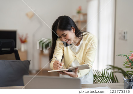 Smiling woman engaged in a phone call while writing in a notebook at her home office. 123775386