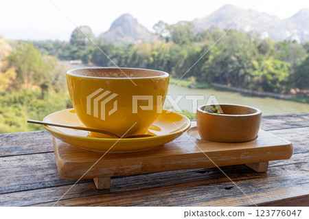Yellow coffee cup and coffee spoon on wooden table with scenic mountain and river view. 123776047