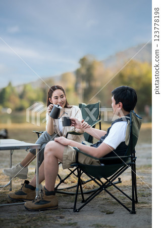 A woman is smiling at the man while pouring drink at folding table, with blurred forest scenery. 123778198