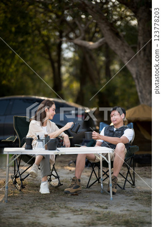 A smiling woman is pouring drink for the man at folding table in front of their camping tent. 123778203
