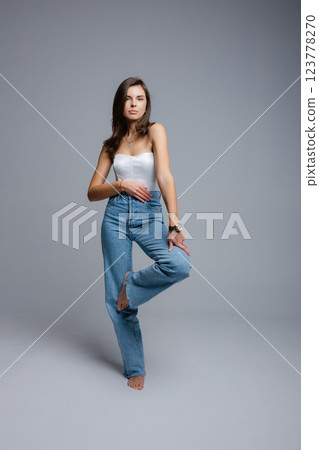 Young woman posing barefoot wearing jeans and white top on gray background Young woman posing barefoot wearing jeans and white top on gray background 123778270