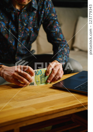 Man holding one hundred euro banknotes, sitting on sofa in front of table with open laptop. Person counting cash money at home. Personal finance and home budget concept Man holding one hundred euro banknotes, sitting on sofa in front of table with open laptop. Person counting cash money at home. Personal finance and home budget concept 123779343