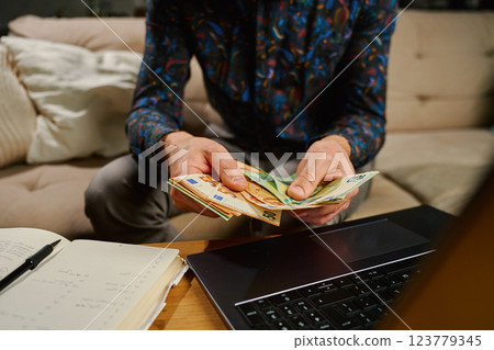 Man holding euro banknotes, sitting on sofa in front of table with laptop and notebook. Person counting remaining money after paying bills and utility expenses. Personal finance and home budget Man holding euro banknotes, sitting on sofa in front of table with laptop and notebook. Person counting remaining money after paying bills and utility expenses. Personal finance and home budget 123779345