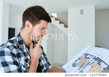 Thoughtful man examines a floor plan while standing in unfinished home. Male home owner analyzing architectural blueprint in new apartment. Interior design and renovation decisions concept 123779364