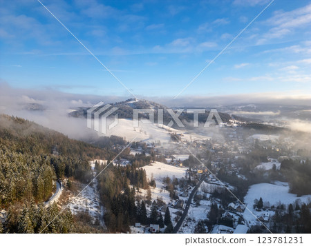 Snow blankets the hills and valleys around Tanvaldsky Spicak, creating a serene winter landscape. Mist hovers low, revealing patches of green and snow under a clear blue sky. 123781231
