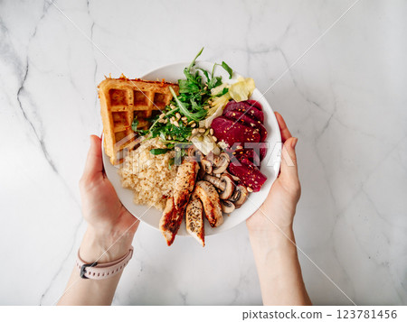Top view of balanced gourmet dish in female hands over white marble tabletop. Bowl with chicken, mushrooms, beetroot, quinoa, green rucola, decorated pine nuts and carrot waffle. Healthy Food concept 123781456