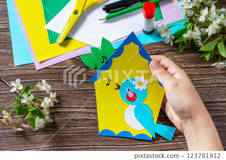 The child holds a spring postcard birdhouse with a bird on a wooden table. Handmade. Project of children's creativity, handicrafts, crafts for kids. 123781912