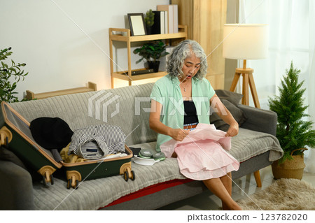 Senior woman sitting on the floor in a living room and packing clothes for vacation 123782020