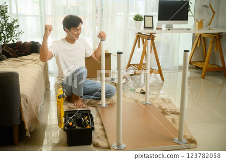 Happy asian man cheering with excitement after successfully assembling a new table at home Happy asian man cheering with excitement after successfully assembling a new table at home 123782058