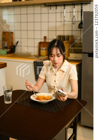 Pretty asian woman enjoys a plate of homemade spaghetti and using smartphone in a cozy kitchen 123782059