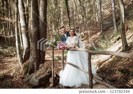 Beautiful bride and an elegant groom standing on forest bridge in nature, surrounded by a trees. 123782394