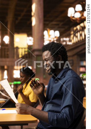 Excited african american student memorizing all the theory from the class lesson, feeling very prepared for his final exams at the campus library. Confident scholar reads through copy books. 123783244