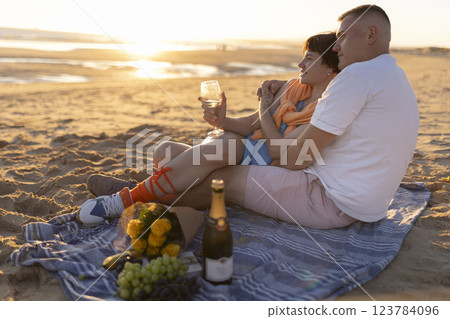 Couple enjoying romantic sunset picnic on beach with wine and flowers Couple enjoying romantic sunset picnic on beach with wine and flowers 123784096