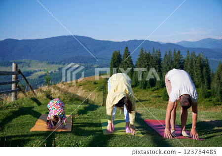 Young family doing yoga outdoors in scenic mountainous landscape in morning. Father, mother and daughter on yoga mats, with scenic view of mountains and forest in background under clear blue sky. 123784485