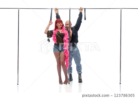 Woman in pink boa standing confidently as man in black leans in behind gripping rail against white studio background. Concept of street style 123786305