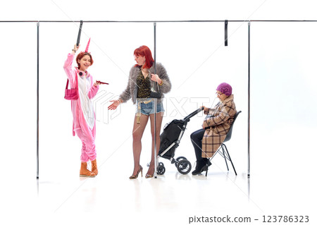 Elderly lady reaching for girl in unicorn onesie standing with phone near stroller, with glamour woman in conversation against white studio background. 123786323