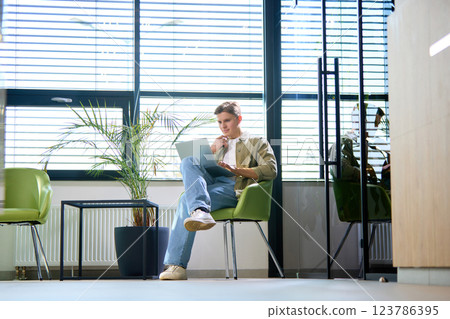 Thoughtful young man, dressed casually, analyzing data on laptop in bright office with contemporary decor and sleek furniture. 123786395