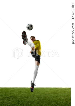 Front view, full-body image of young man, soccer player kicking ball in motion in a jump, with outstretched leg against white background. Front view, full-body image of young man, soccer player kicking ball in motion in a jump, with outstretched leg against white background. 123786409