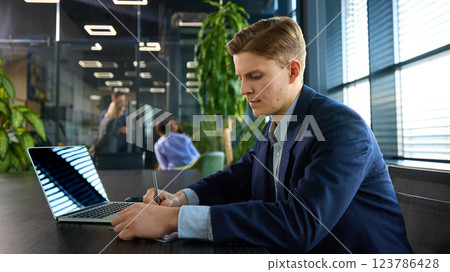 Attentive young businessman writing in notebook at desk with laptop open, maintaining structured approach in professional setting. 123786428